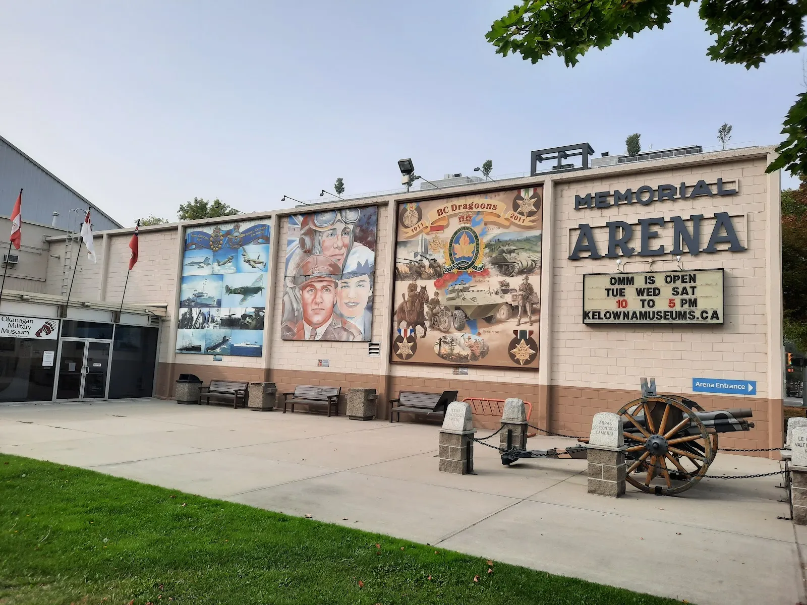 Vista a nivel de la calle de la entrada de Memorial Arena, con grandes letras de 'MEMORIAL ARENA' en la pared, un letrero indicador de 'Arena Entrance', murales conmemorativos de guerra y la entrada compartida del Okanagan Military Museum. — Kelowna Memorial Arena