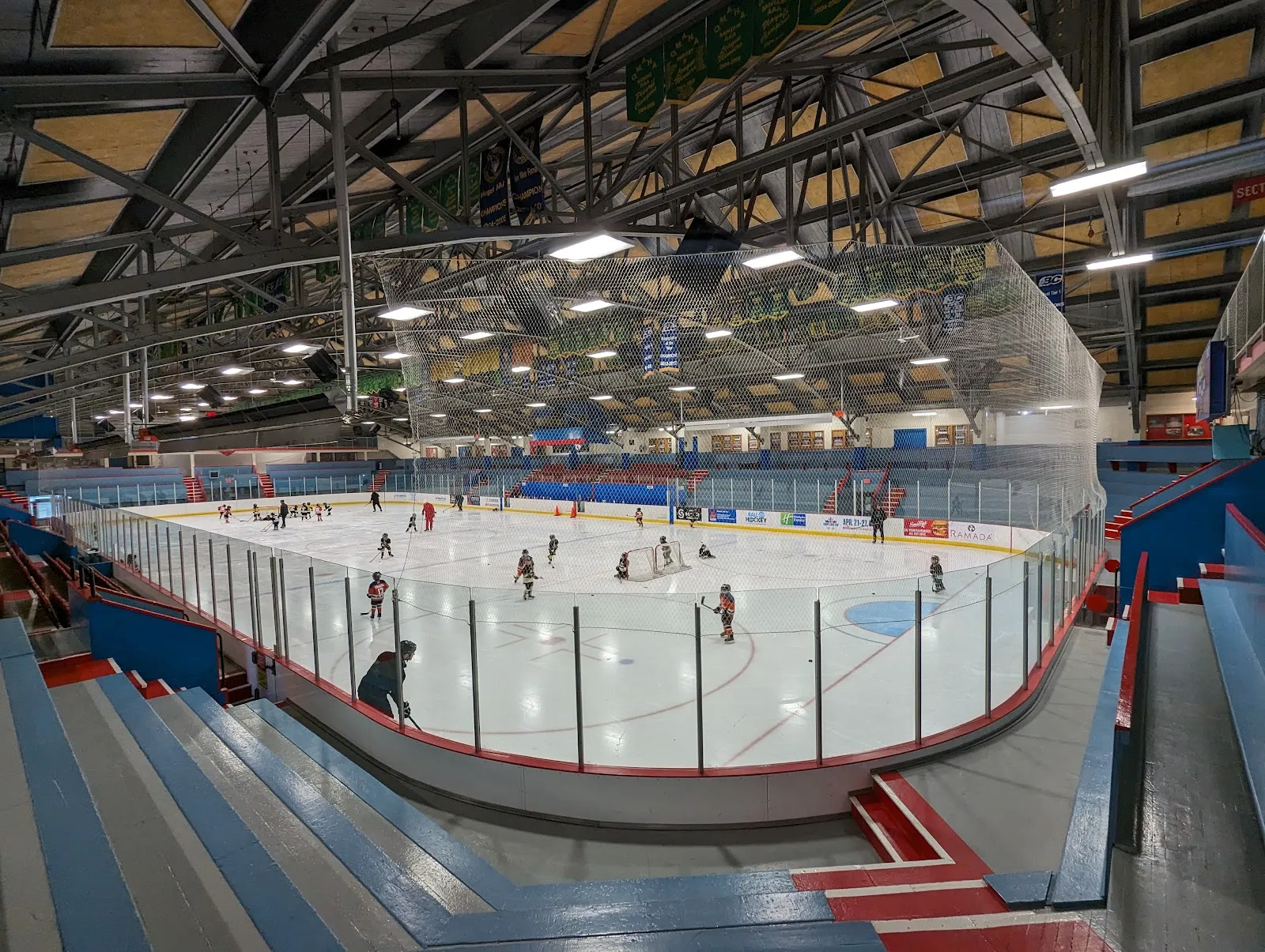 Amplia vista interior de toda la pista desde las gradas, con las vallas, mantas de campeonato arriba y un partido de hockey menor en curso. — Kelowna Memorial Arena