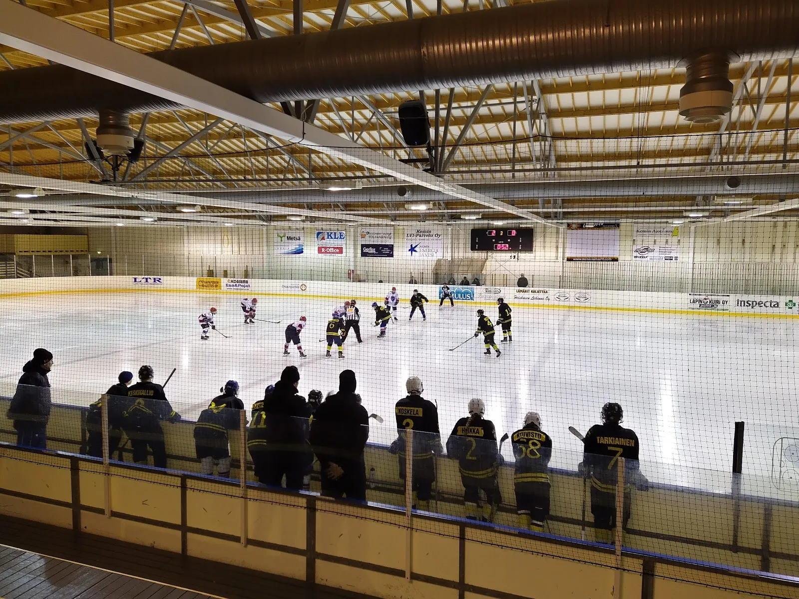 Spectators silhouetted against the lit ice at the Kemi ice hall with skaters across the rink and the timber ceiling above. – Kemin jäähalli
