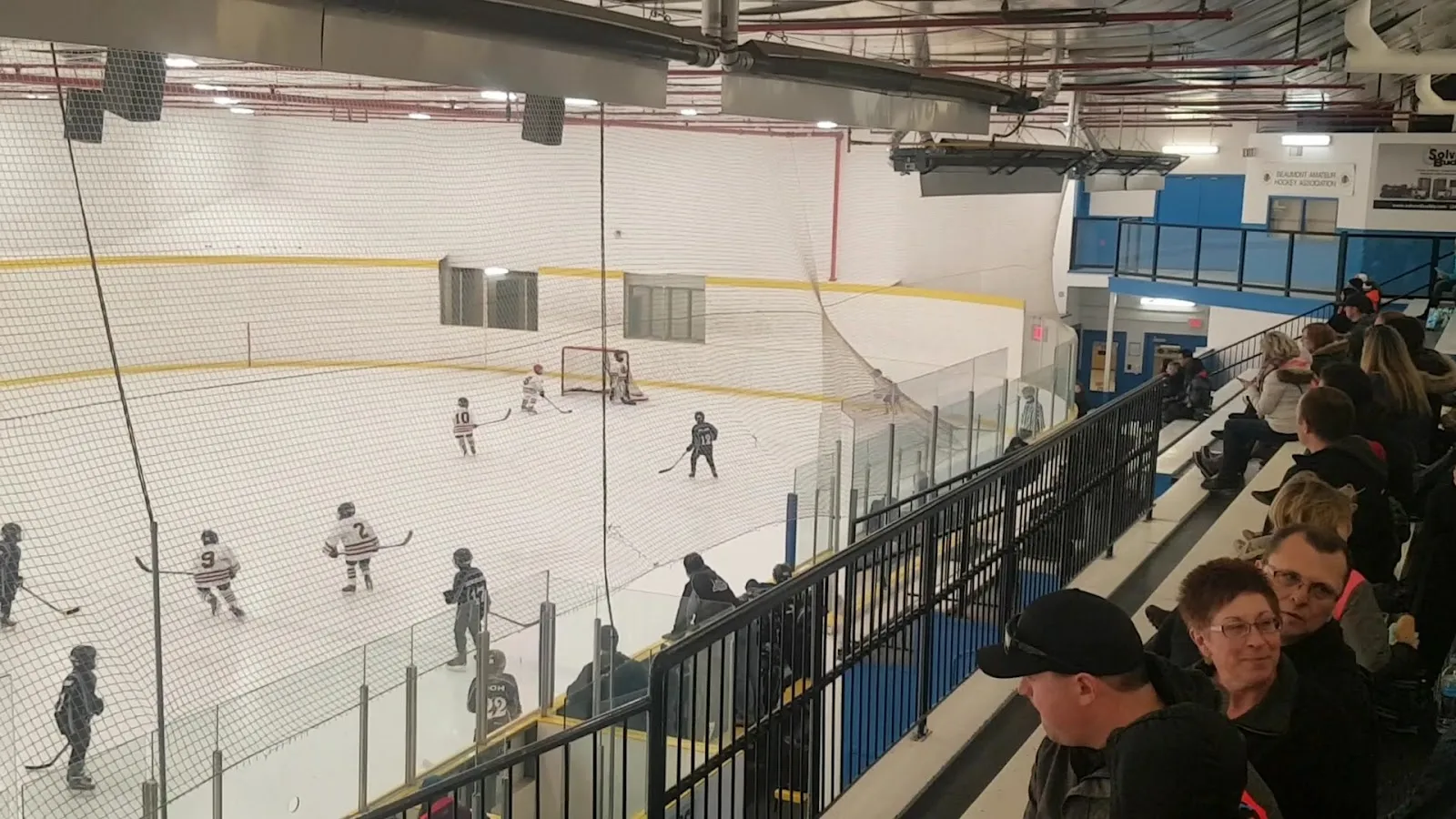 A game seen along a railed spectator bench, players spread across the sheet under a white insulated ceiling with glass panels along the near side. — Ken Nichol Regional Recreation Centre