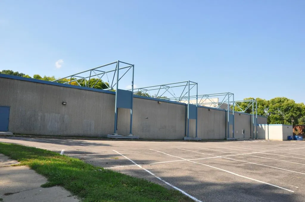El largo muro lateral de concreto de la arena con sus armazones de techo, del otro lado de un estacionamiento vacío. — Ken Yackel-West Side Arena