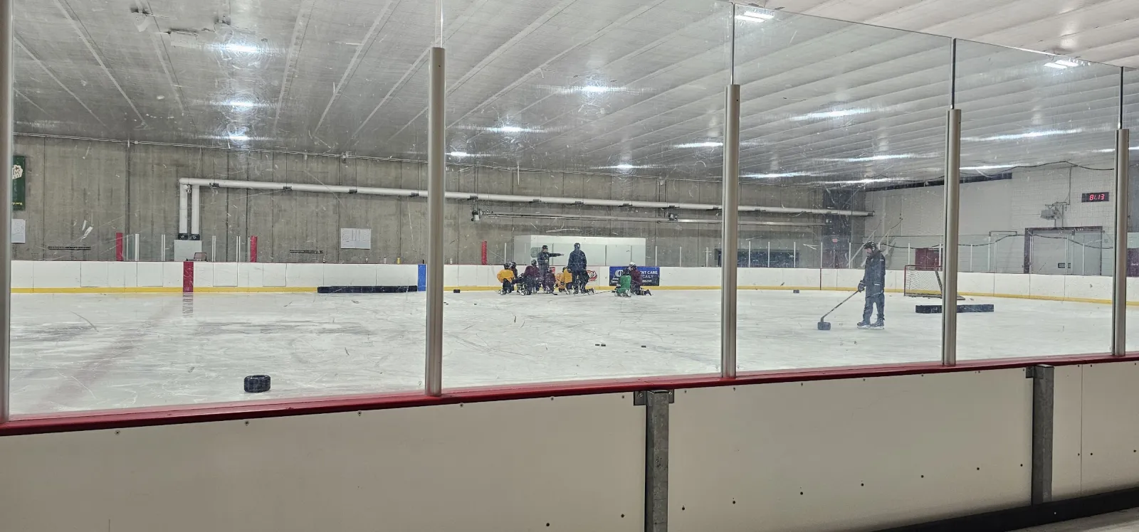El hielo visto a través del vidrio desde el lado de las bancas, con algunos jugadores y pucks cerca de las bardas del fondo bajo un techo bajo y blanco. — Ken Yackel-West Side Arena