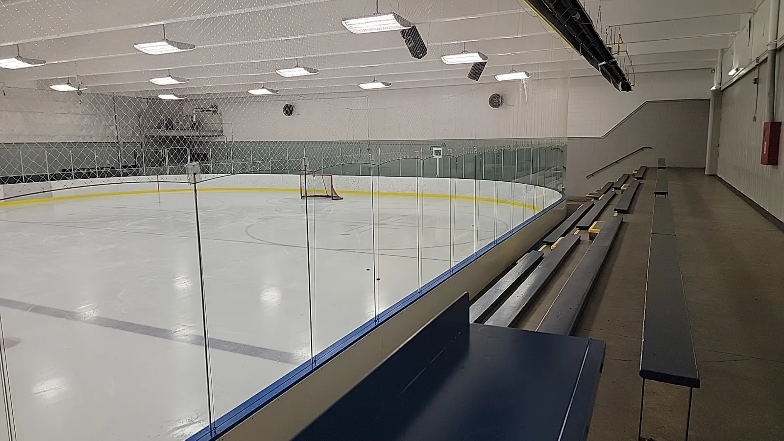 The empty ice seen from the benches behind the glass, with the far boards, a Canadian flag and sponsor banners under a white ceiling. — Kenilworth Arena
