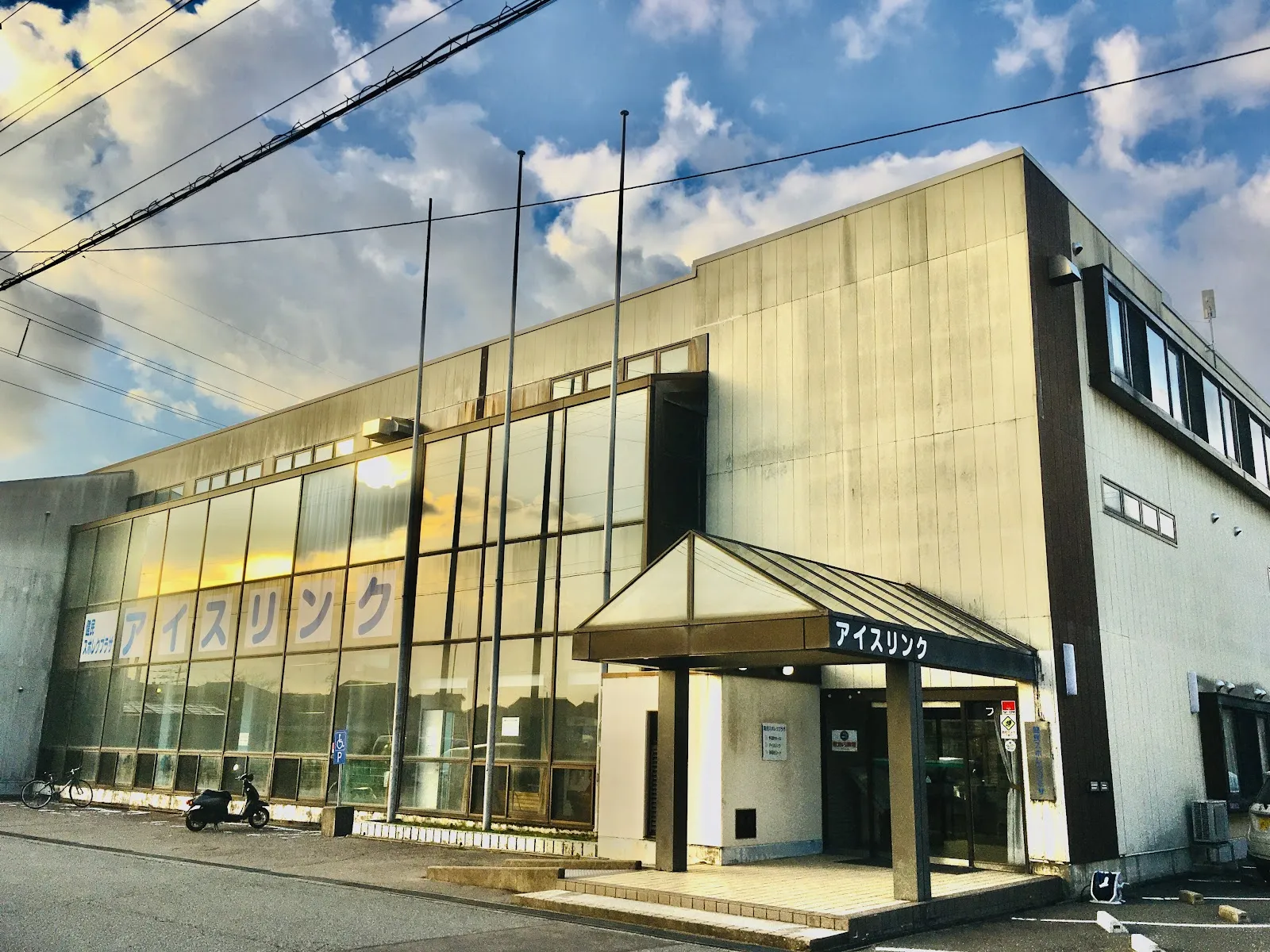 夕暮れの健民スポレクプラザのガラス張りの建物。上の階の窓に「アイスリンク」の看板があり、下に庇のある入口があります。 — Kenmin Sporec Plaza Ice Rink