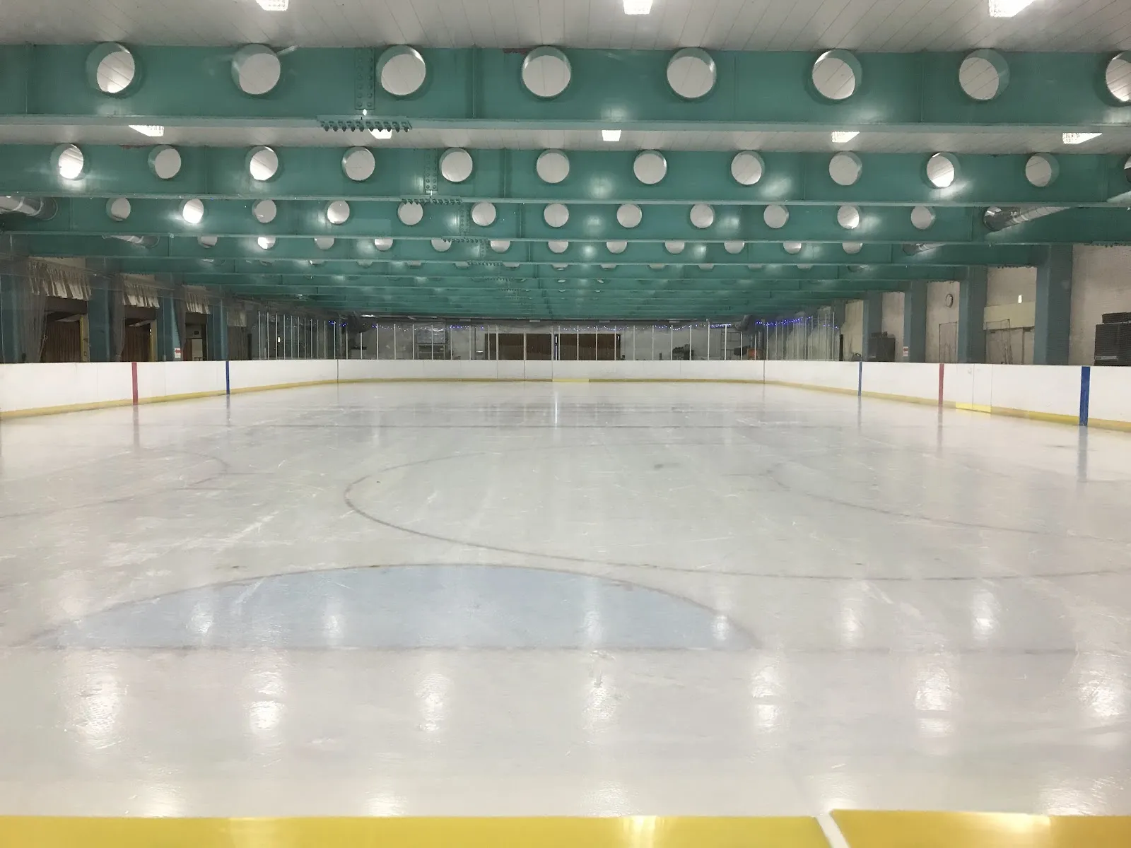 長い青緑色の天井に丸い照明が並ぶ、健民スポレクプラザのリンクの空の氷面。センターサークルと黄色いフェンスの上端が見えます。 — Kenmin Sporec Plaza Ice Rink