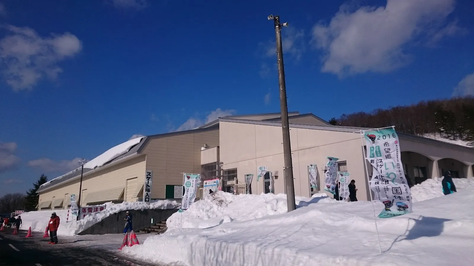 冬の青少年の家の細長い低層の建物。正面に沿って雪が積み上げられ、脇に電柱が立っています。 — Kenpoku Youth House Skating Rink (Stellapal)