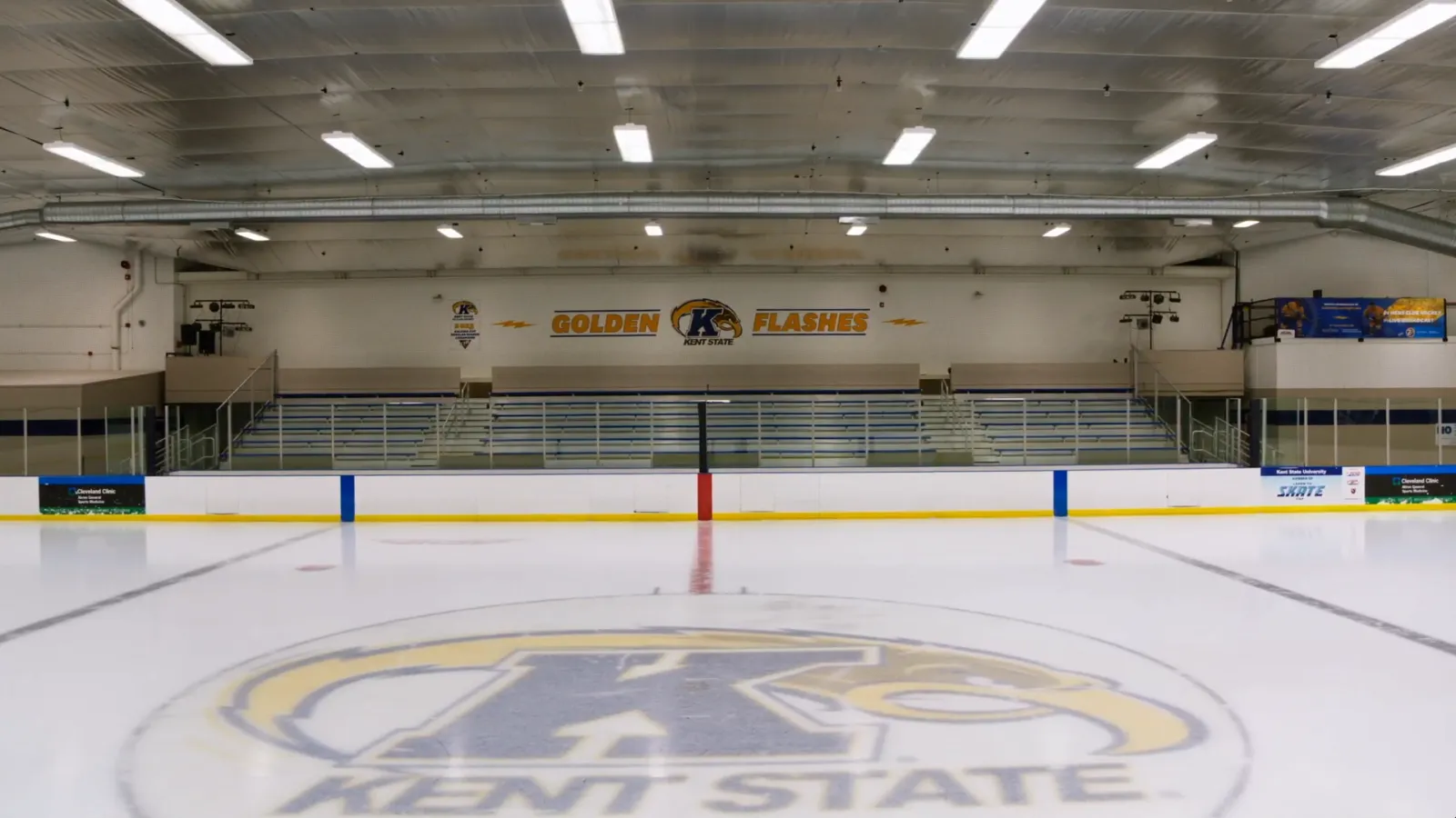 Empty rink at ice level facing the spectator bleachers, with a Kent State "Golden Flashes" wall graphic above the stands and the Kent State Golden Flashes logo painted at center ice. — Kent State Ice Arena