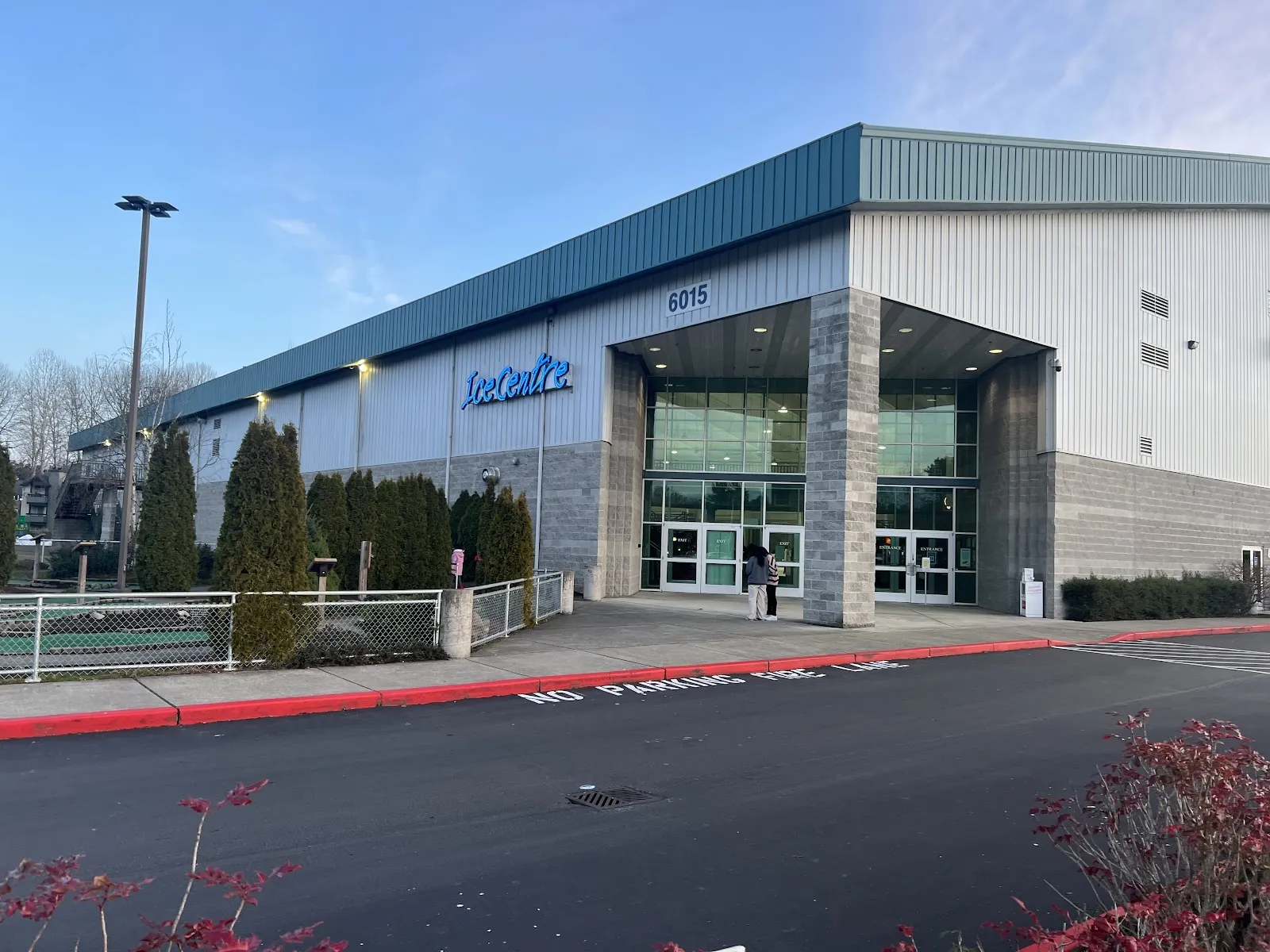 Front entrance of Kent Valley Ice Centre, a metal-panel building with a blue "IceCentre" wall sign above stone-clad entry columns, evergreen shrubs, a mini-golf course to the left, and the parking lot in the foreground — Kent Valley Ice Centre