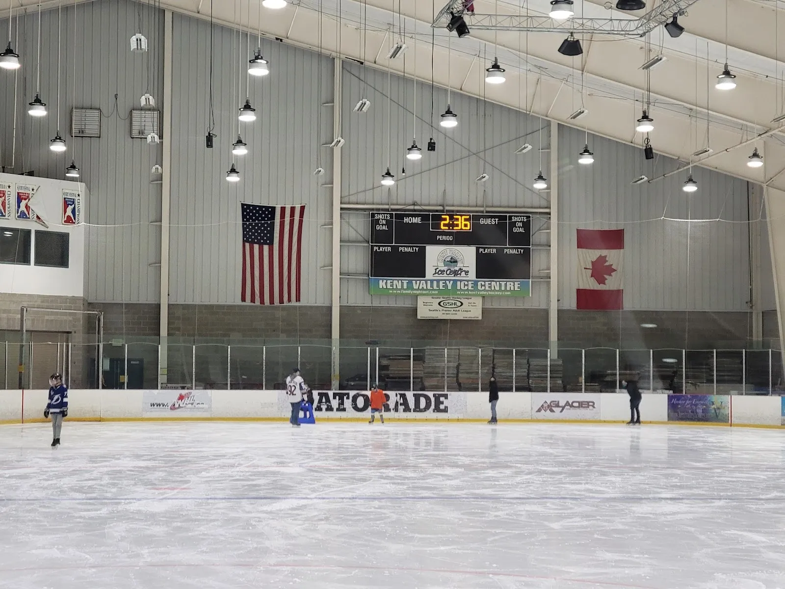 A long view across the sheet with American and Canadian flags and a scoreboard on the far wall, a lone player at the left edge under a steel-trussed roof. — Kent Valley Ice Centre