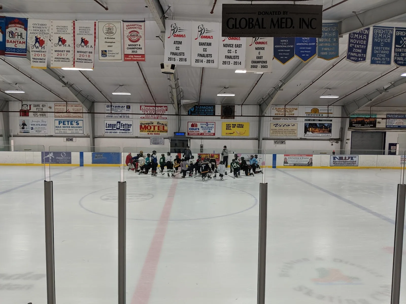 Un equipo reunido en el centro del hielo en el extremo lejano de una pista amplia detrás de una hilera de postes de vidrio, con banderines de patrocinadores sobre las vallas. — King Edward Park Arena (Brighton)
