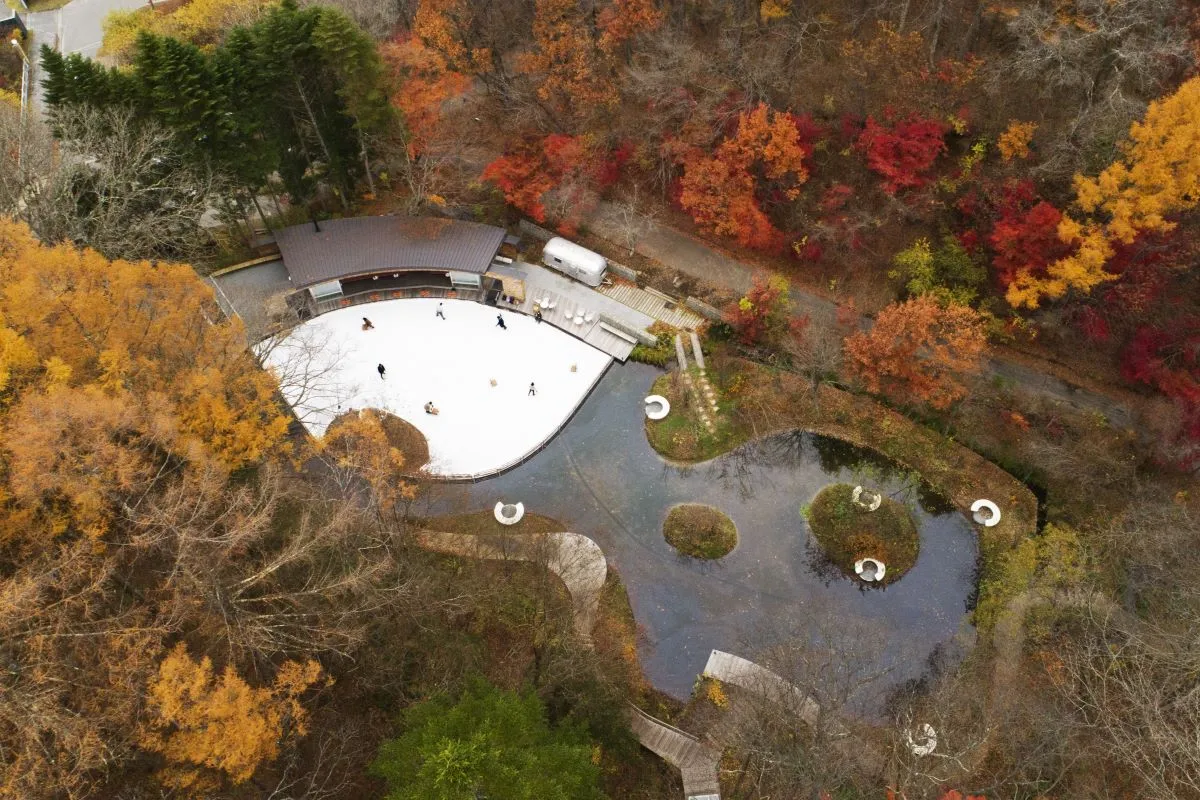 紅葉のカエデとカラマツに囲まれたケラ池を空から見た写真。白い氷のそばに曲線を描くカフェの建物と、奥に暗い池が見える。 — Kera-Ike Skate Rink