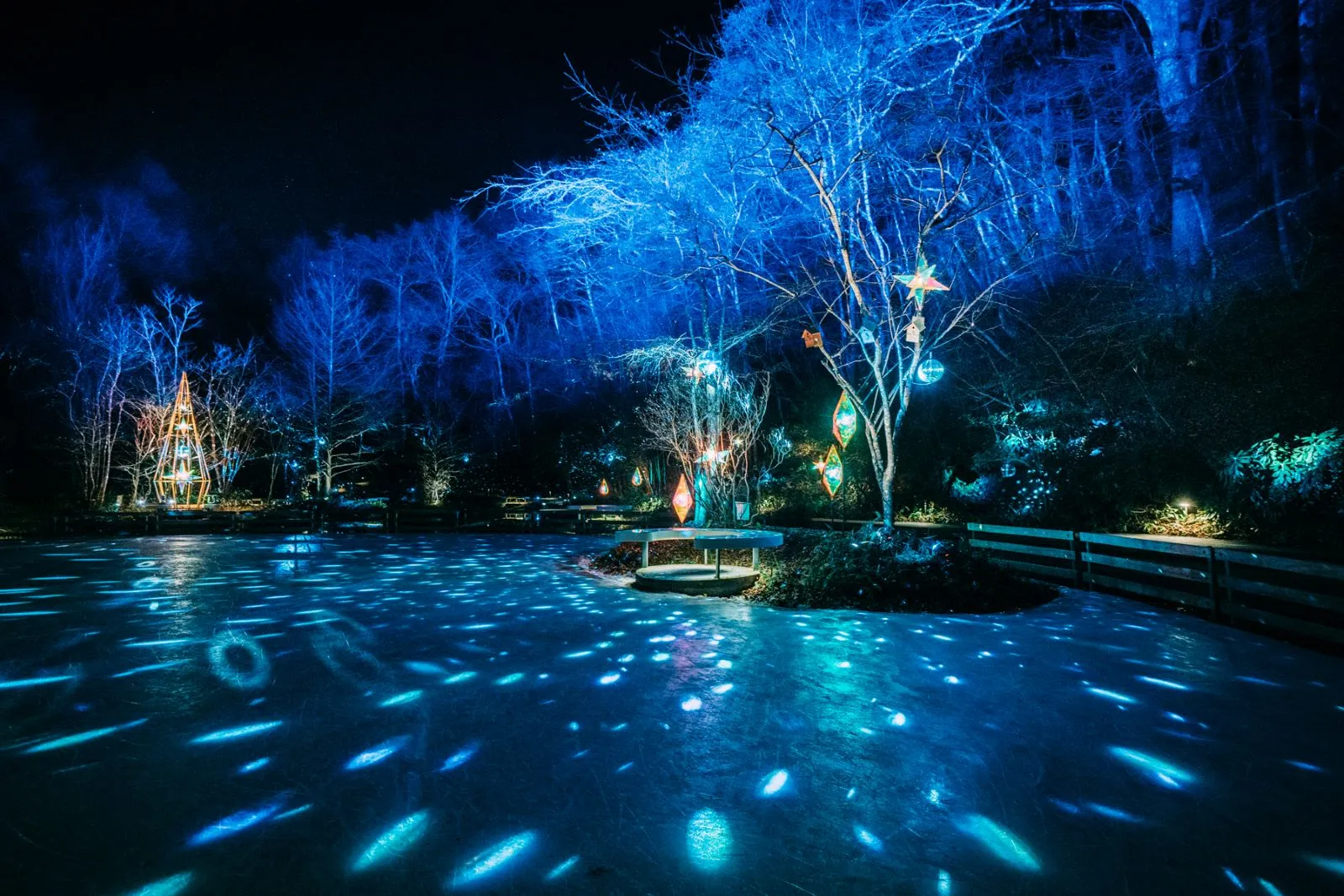 冬の「星屑」イルミネーションで夜に青く照らされたケラ池の氷。葉の落ちた木々とランタンが森の池を囲んでいる。 — Kera-Ike Skate Rink