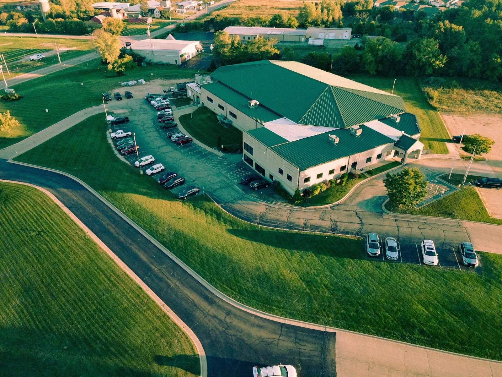 El Kettle Moraine Ice Center desde el aire al atardecer: un edificio verde de la pista con techo a cuatro aguas, un ala frontal más baja y estacionamiento a lo largo de un camino de acceso curvo. — Kettle Moraine Ice Center