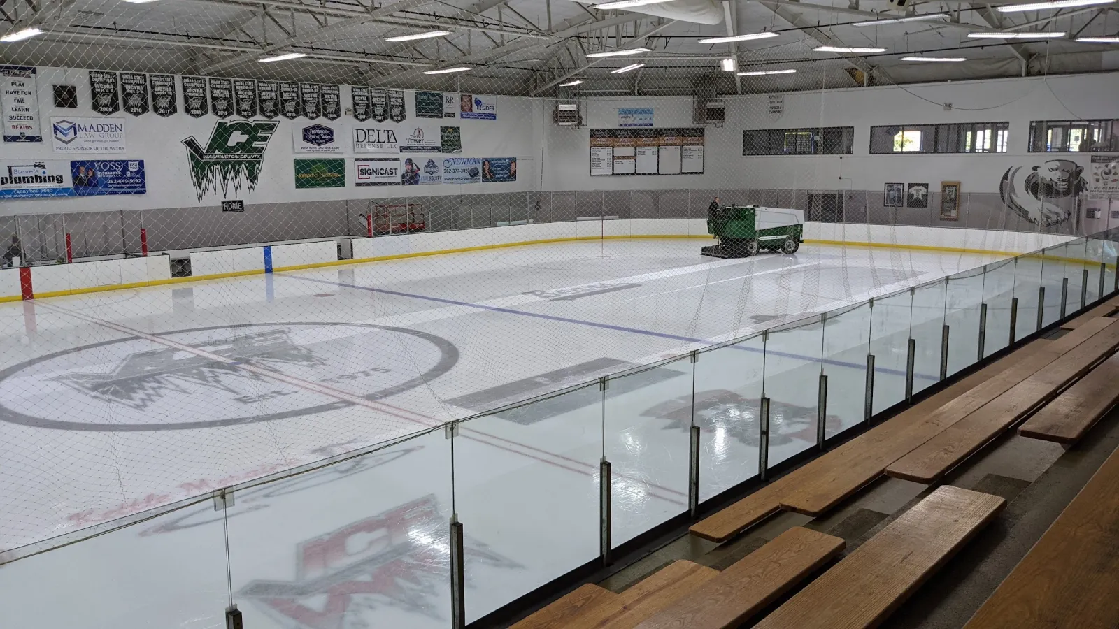 El hielo visto desde las gradas, con una pulidora de hielo en el extremo opuesto, el logotipo del centro de la pista y mantas de patrocinadores alrededor de la pista. — Kettle Moraine Ice Center