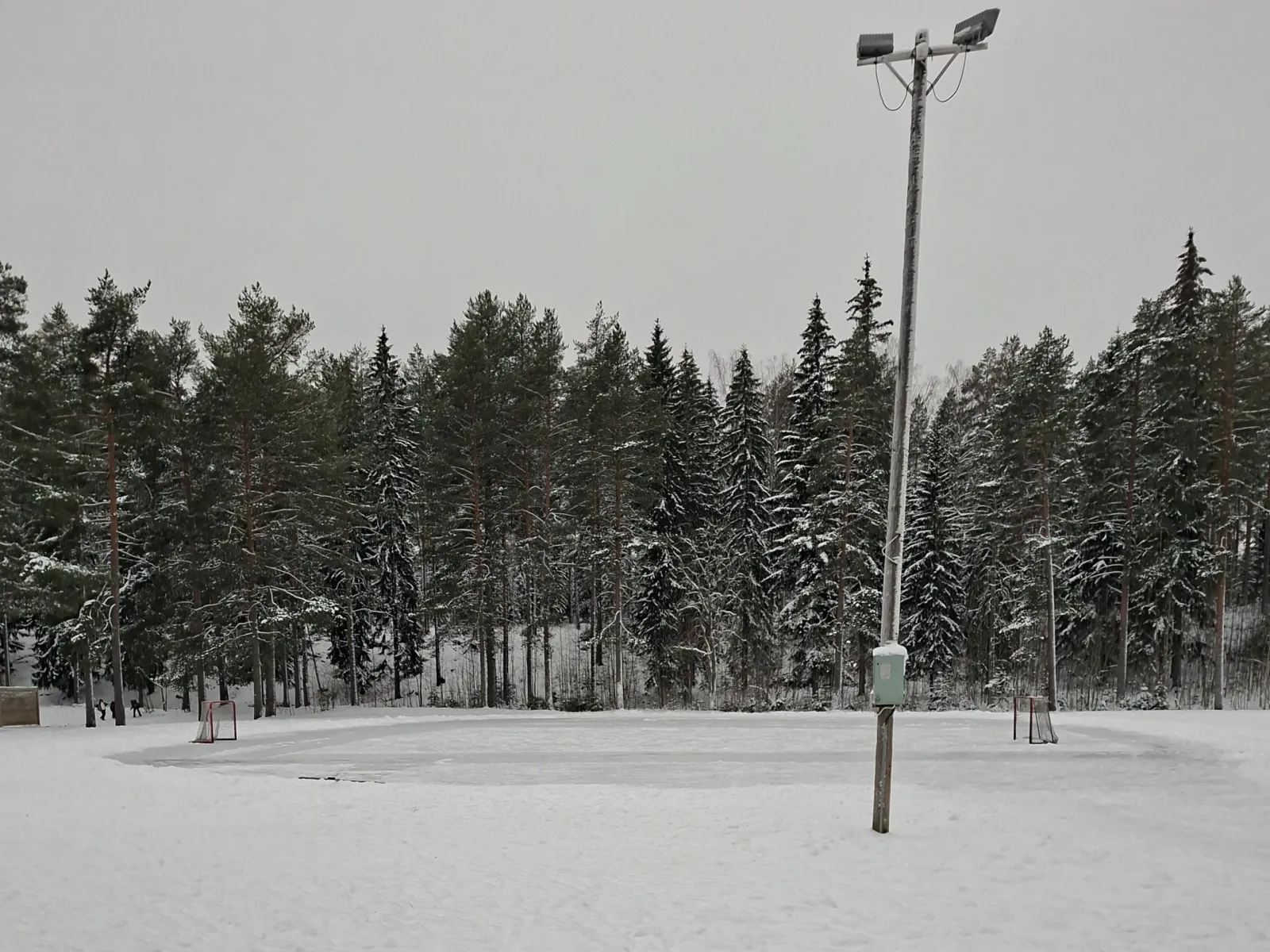 The Kettulanlahti school skating field in winter, with a floodlight pole and a goal at the far end among the pines. — Kettulanlahden koulun luistelukenttä