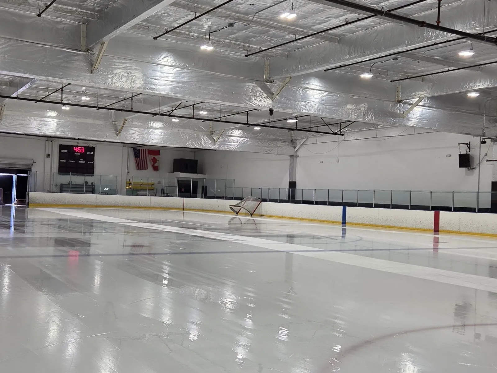 Hielo recién pulido y vacío en KHS ICE a nivel de la pista, con una portería empujada hacia las vallas del fondo bajo un techo aislado plateado y el marcador — KHS ICE