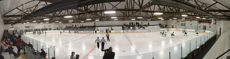 A panorama of the Killam Memorial Arena ice during a hockey game, under the arched wooden roof trusses, with spectators along the glass. — Killam Memorial Arena