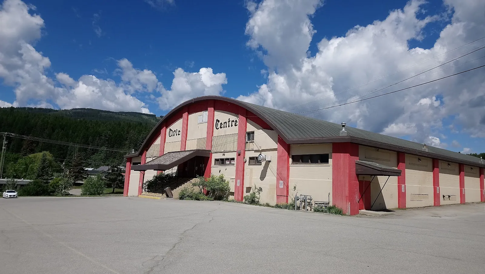El Kimberley Civic Centre, con tejado en arco, nervios rojos y paredes crema, y su nombre en letra gótica sobre el hastial, visto desde el aparcamiento con colinas boscosas y grandes nubes blancas detrás. — Kimberley Civic Centre Arena