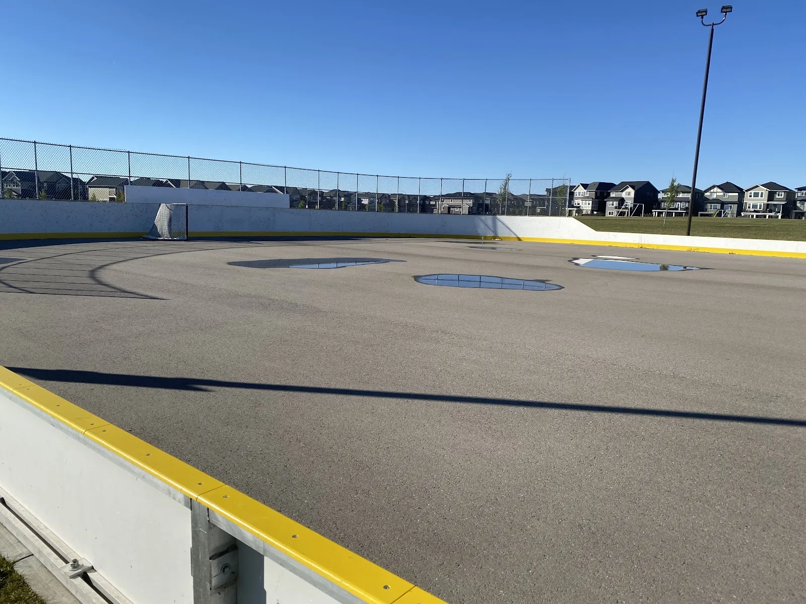 The boarded outdoor rink at Kings Heights in summer: white boards with a yellow cap, fence, floodlight pole and the surrounding houses. — Kings Heights Drive Boarded Rink (Airdrie)