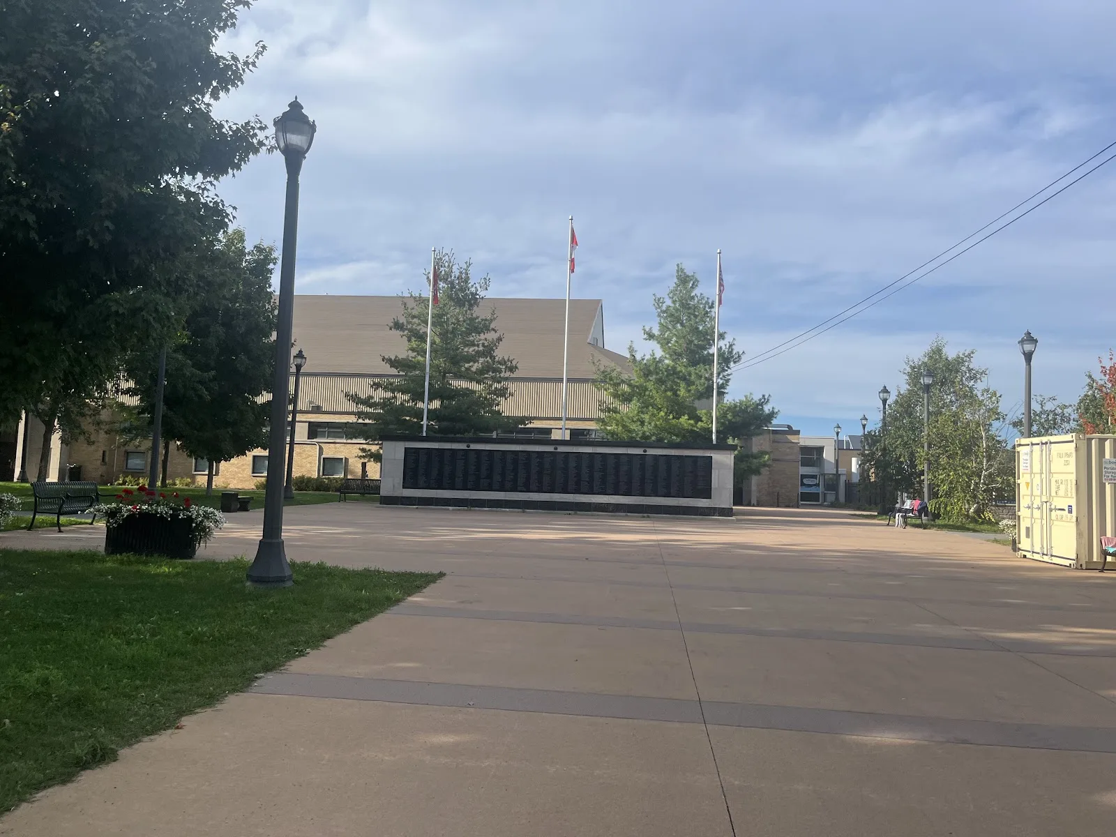 The Memorial Centre's tan brick front beyond the Memorial Wall, with flagpoles and a lamp-lined walkway through the grounds. — Kingston Memorial Centre
