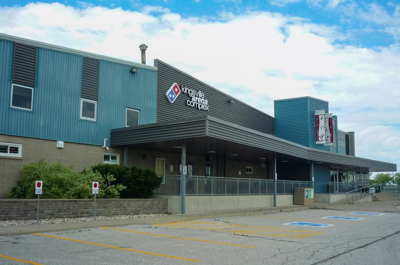 Kingsville Arena Complex: a blue-clad arena block with a dark entrance canopy and the complex sign above, seen from the accessible parking spaces in front. — Kingsville Arena (Kingsville Recreation Complex)