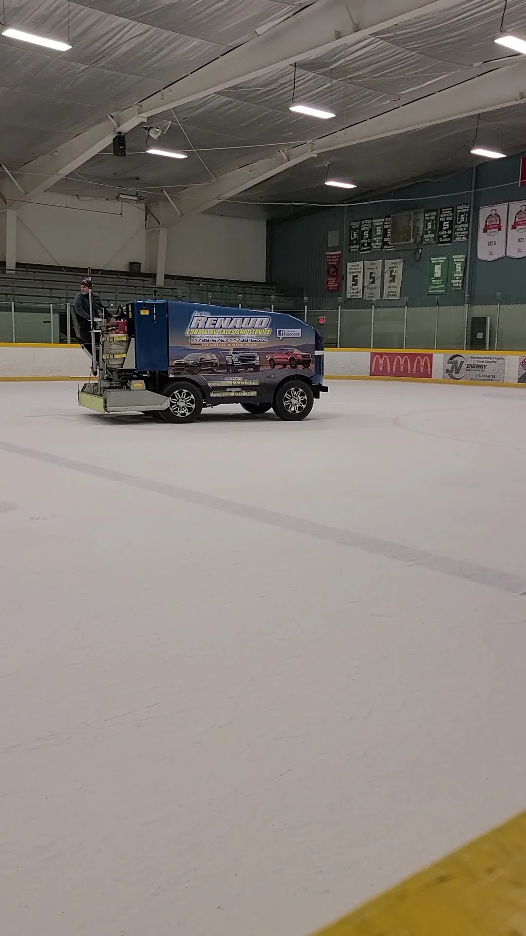 Fresh ice at Kingsville Arena with the resurfacer at the far end under the steel roof and banners — Kingsville Arena (Kingsville Recreation Complex)