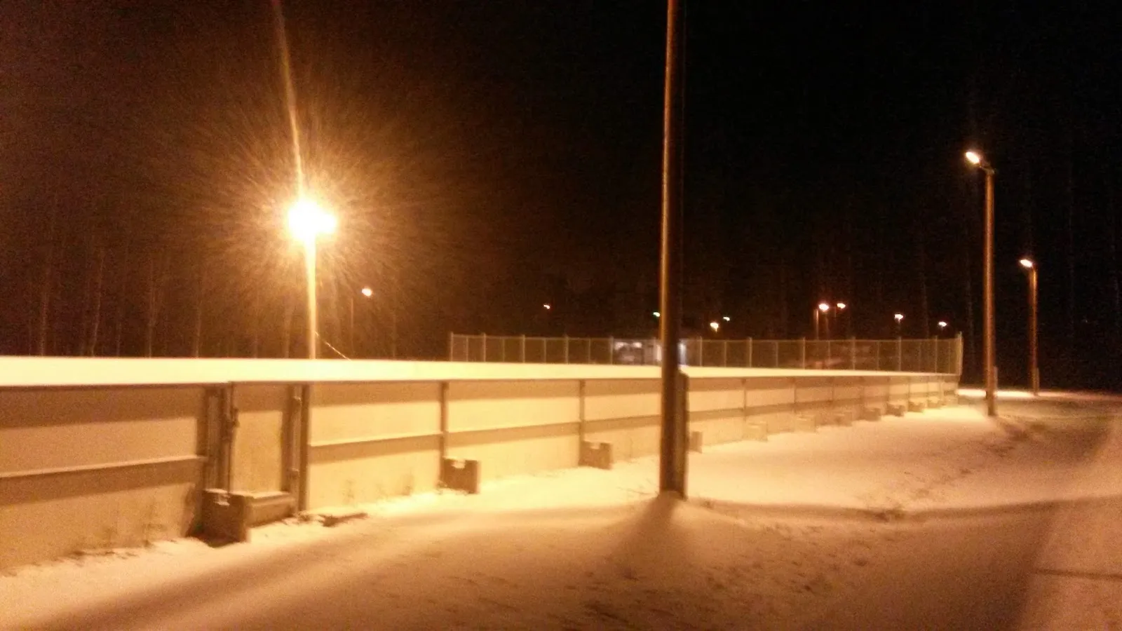 The boards of the floodlit outdoor rink at Kinkomaa on a snowy night, with street lamps along the path beside it – Kinkomaan kaukalo