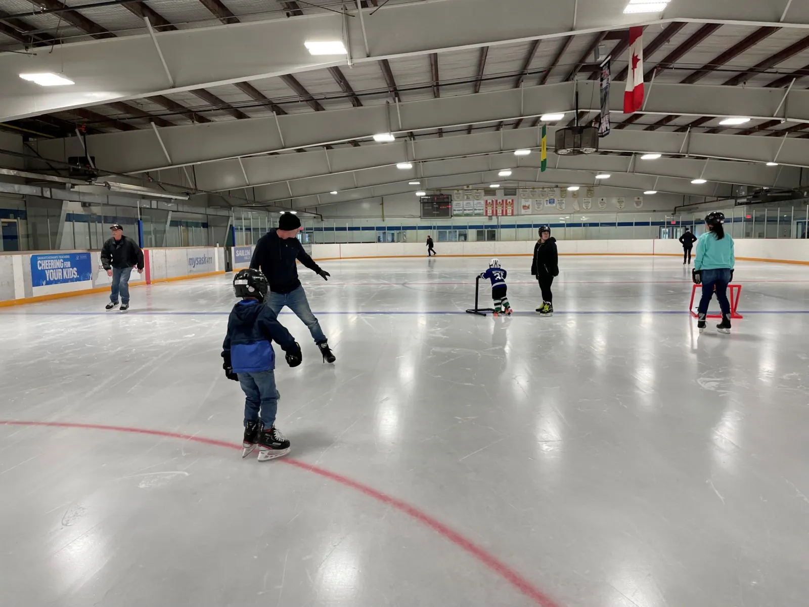 Skaters and families spread across a wide sheet in a public session, a bright white surface under a steel-trussed roof with Canadian and provincial flags on the far wall. — Kinsmen Arena (Moose Jaw)