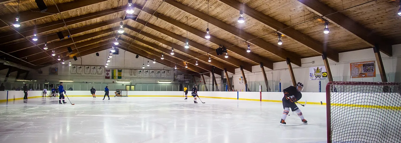The ice at Kinsmen Arena under its timber-beamed roof, seen from the far end with a single player near the net — Kinsmen Arena (Prince Albert)