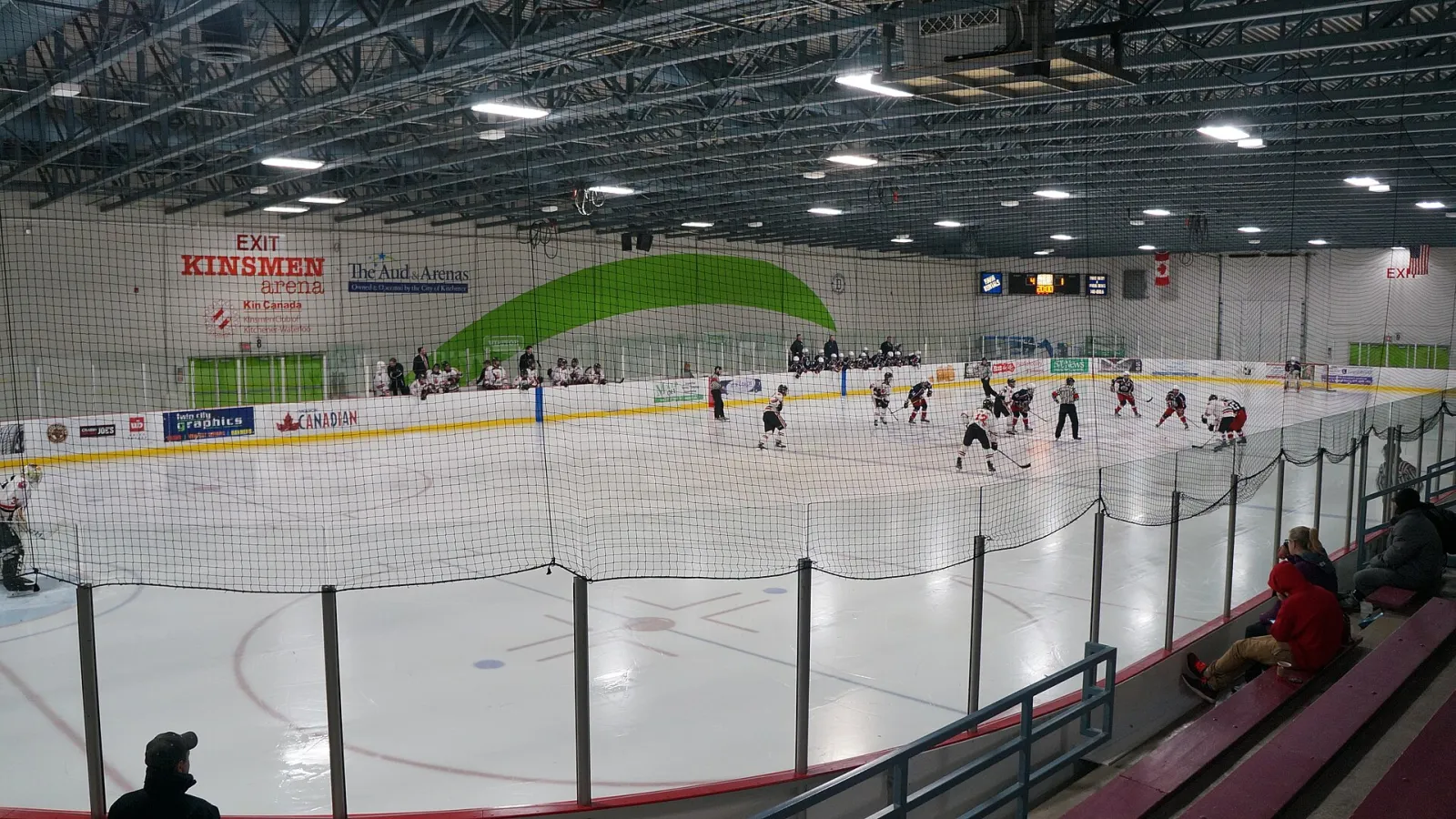 El hielo del Kinsmen Arena con sus vallas rojas y blancas, el nombre Kinsmen en el muro del fondo y un partido en marcha en el extremo lejano. — Kinsmen Arena (The Aud)