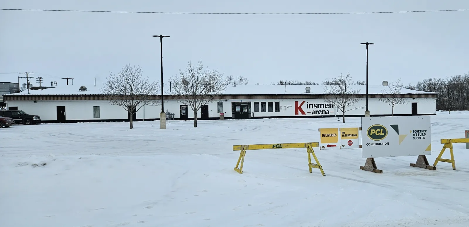The long white Kinsmen Arena building with its red-and-black Kinsmen Arena lettering, seen across a snowy lot in January 2025, with the renovation contractor's signs and barricades in front. — Kinsmen Arena (Yorkton)