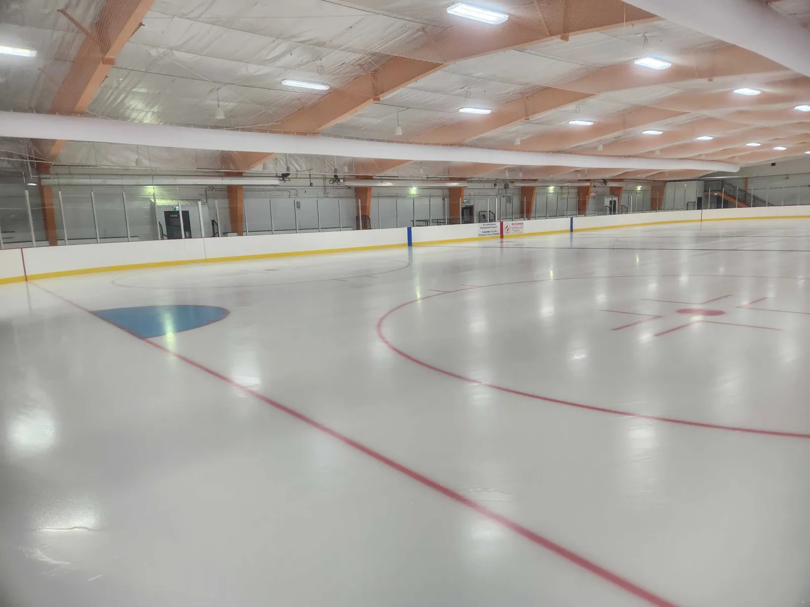 The rebuilt Kinsmen Arena ice from the corner: new white boards and glass, red and blue lines, and exposed orange roof beams under LED lights. — Kinsmen Arena (Yorkton)