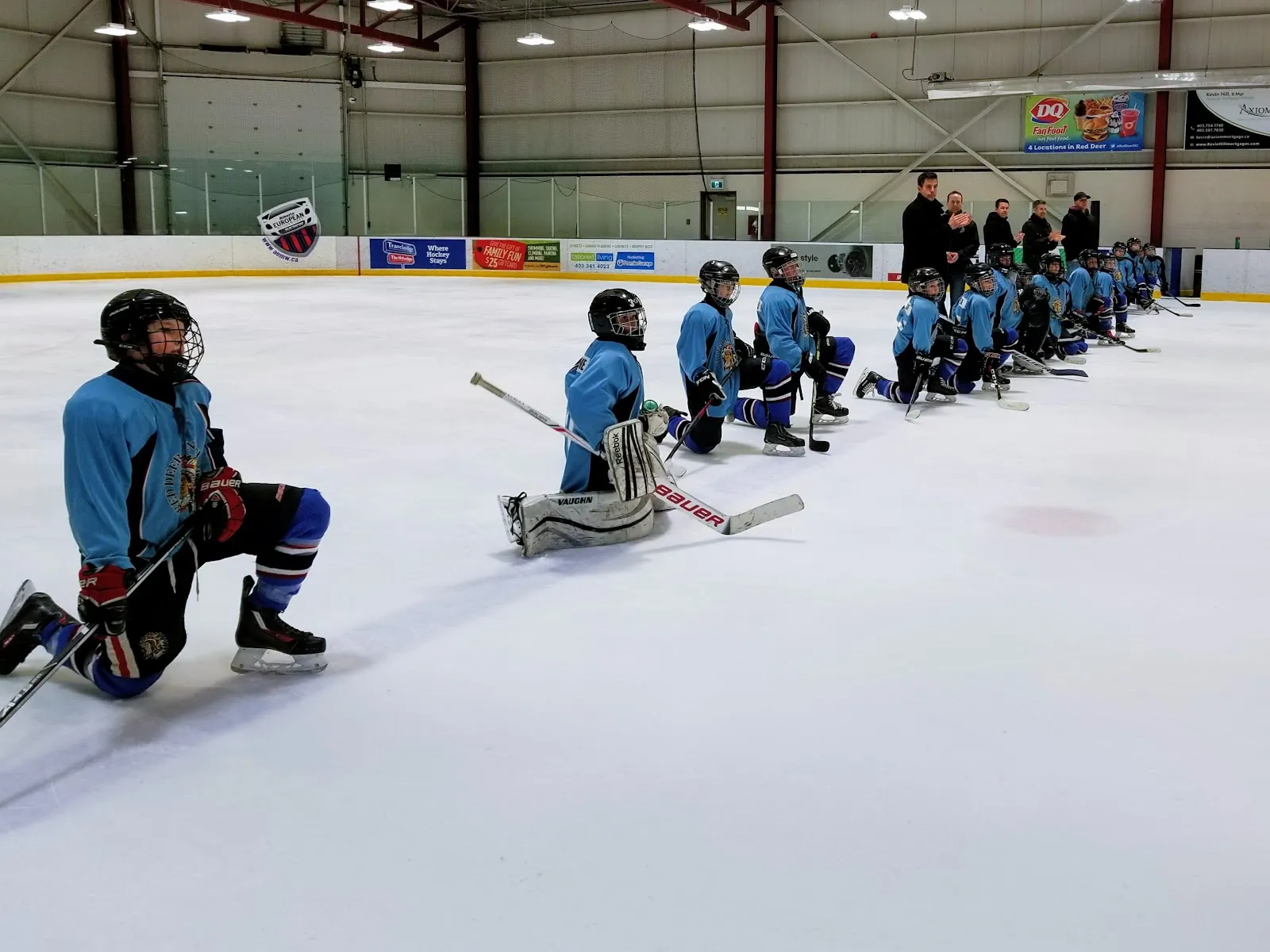 El amplio interior de la arena con la pista de hielo y una práctica de hockey juvenil en el extremo opuesto, vista desde las vallas a la distancia. — Kinsmen Community Arenas