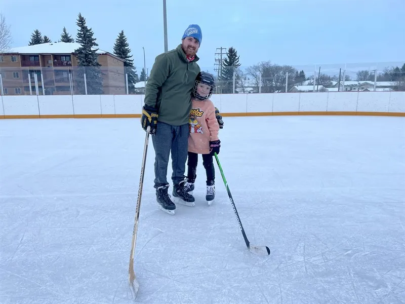 A hockey game on the ice of an outdoor rink, with players around the net and spectators watching behind the boards and fence. — Kinsmen/Scott Seaman Outdoor Sports Rink (Ponoka)