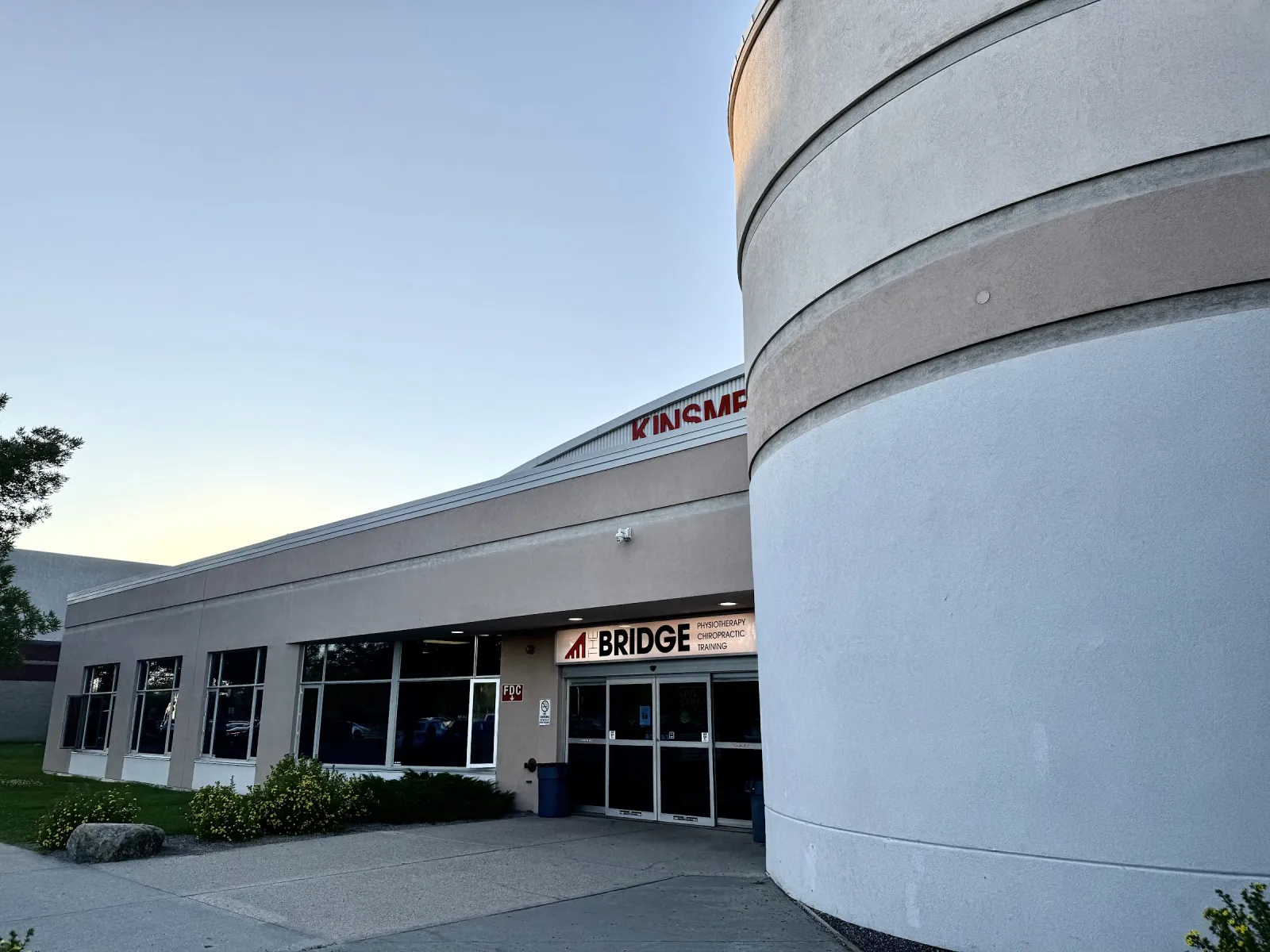 The Kinsmen complex entrance at dusk, with a curved grey wall, glass doors and a Bridge clinic sign on the long low building. — Kinsmen Twin Arena
