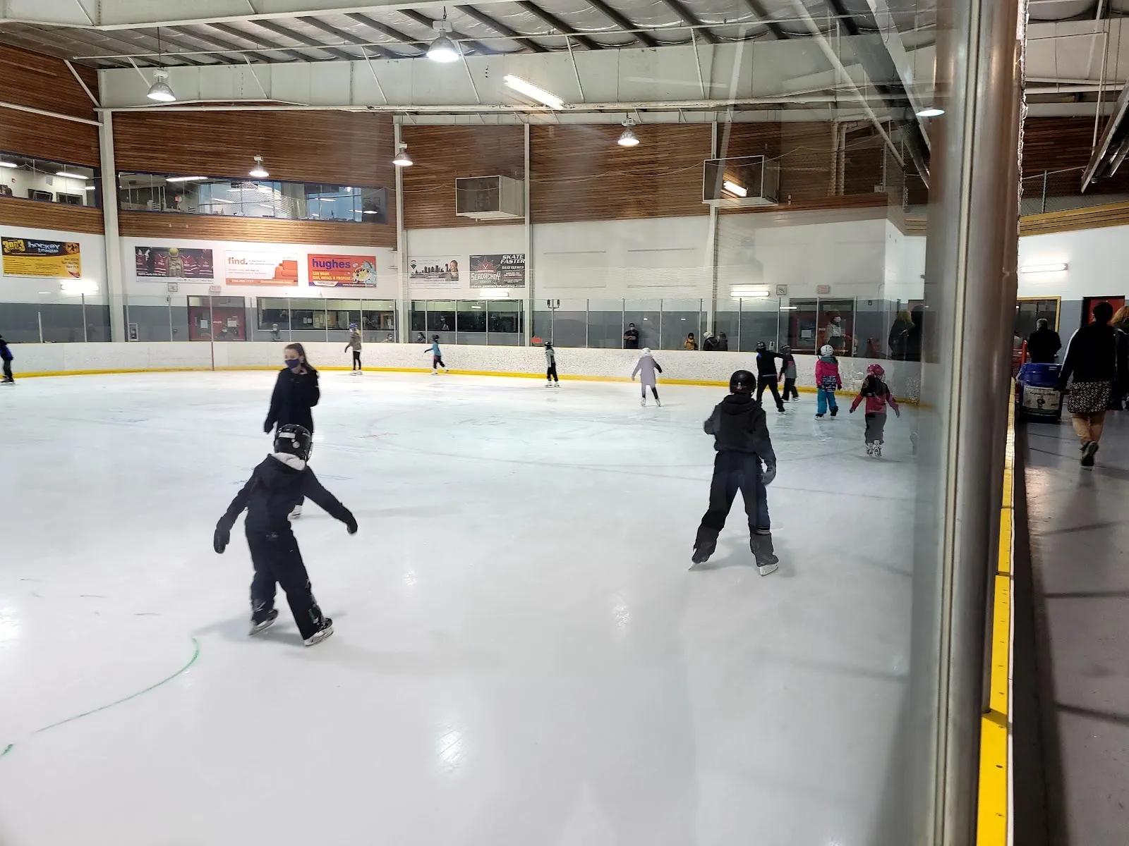 A public skating session on a wide sheet, skaters spread across the ice below a timber-lined wall with a long band of windows above the boards. — Kinsmen Twin Arena