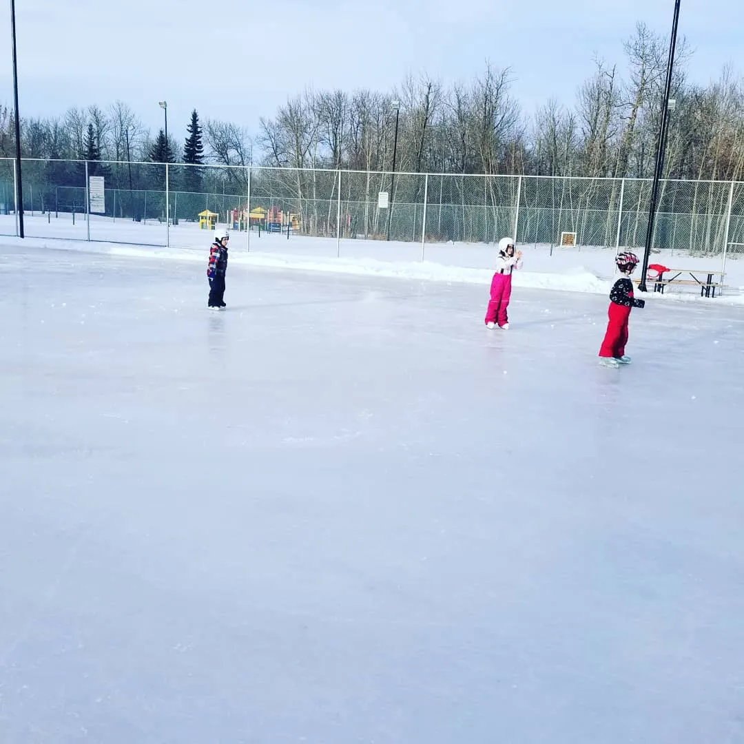 Children skating on the fenced outdoor rink at Kinsmen/Westboro Park in Sherwood Park, with a playground and picnic table behind the fence — Kinsmen/Westboro Park Boarded Rink