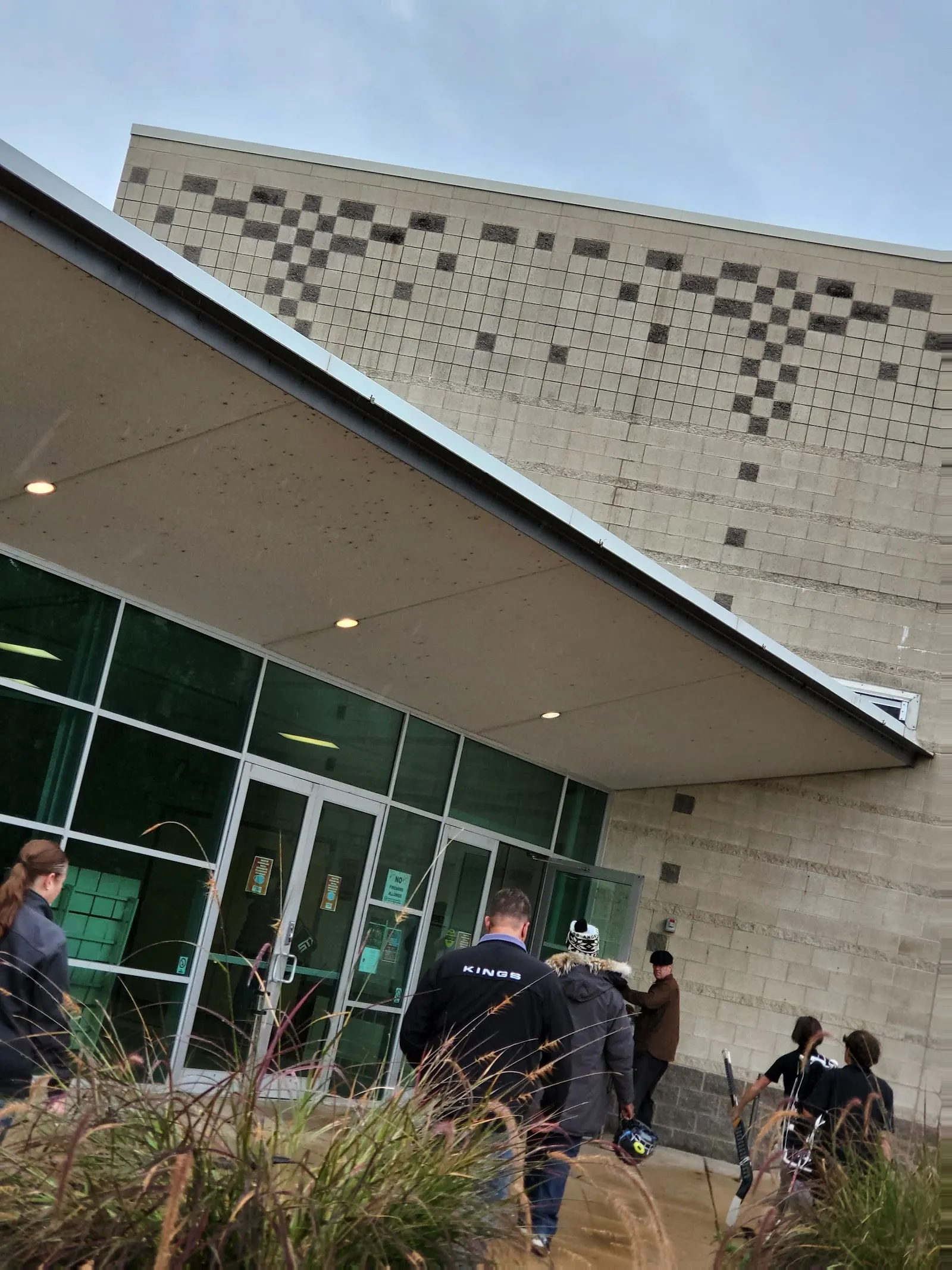 The glass entrance doors of the Kirkwood Ice Rink beneath a flat canopy, a pale-brick wall with a logo beside them and a green-tinted glazed facade. — Kirkwood Ice Rink