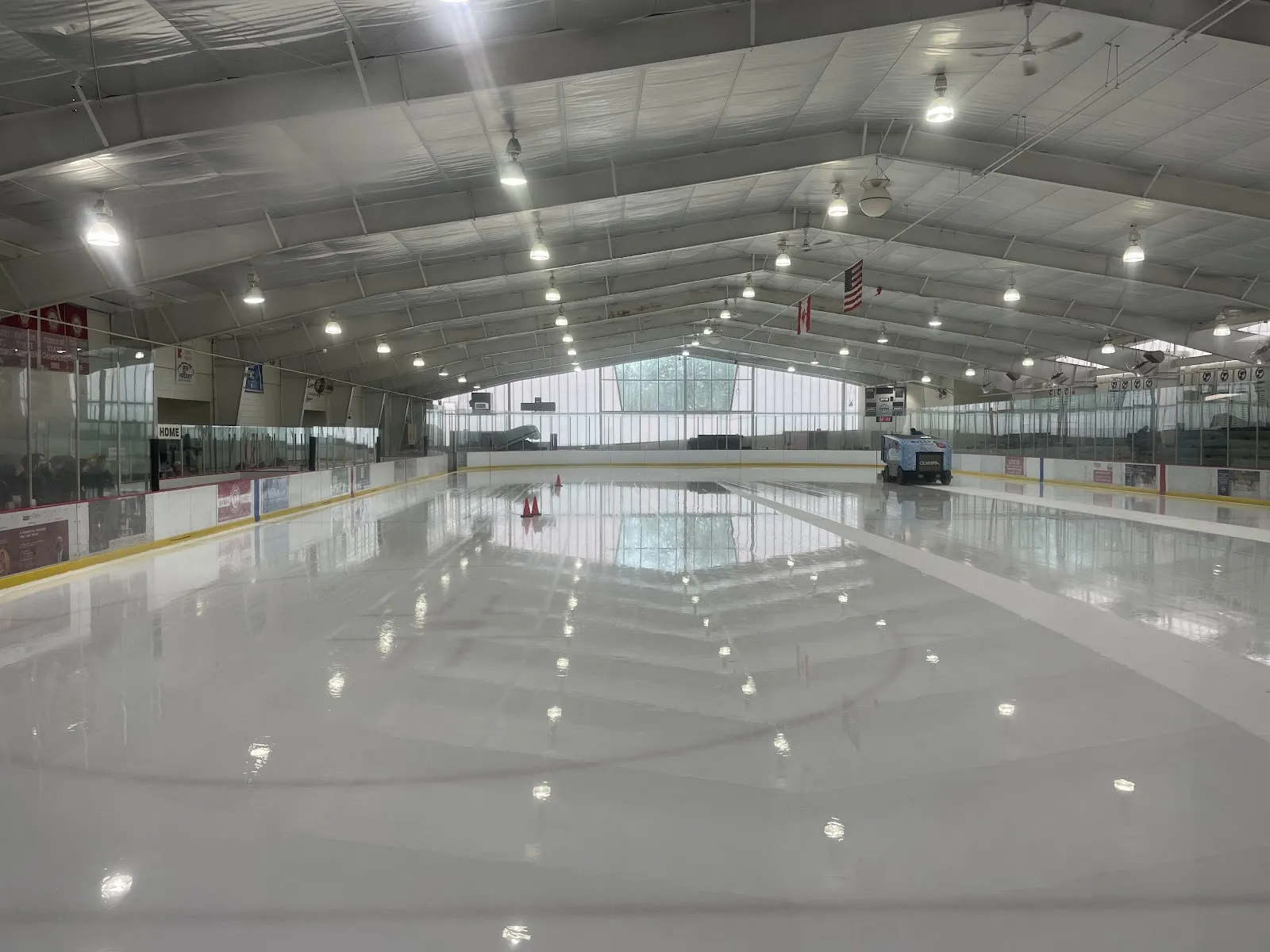 Freshly resurfaced, empty ice sheet from the boards: a Zamboni-style resurfacer parked at the far end, American and Canadian flags hanging from the arched roof trusses, and a bank of windows across the back wall. No people on the ice. — Kirkwood Ice Rink