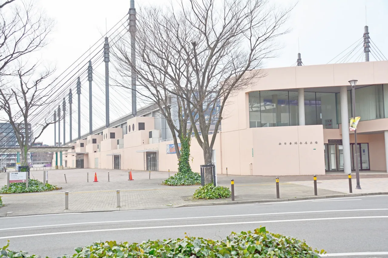 冬枯れの木々越しに見た北九州アイススケートセンターの明るい色の建物。背後に鉄塔がそびえています。 — Kitakyushu Ice Skate Center