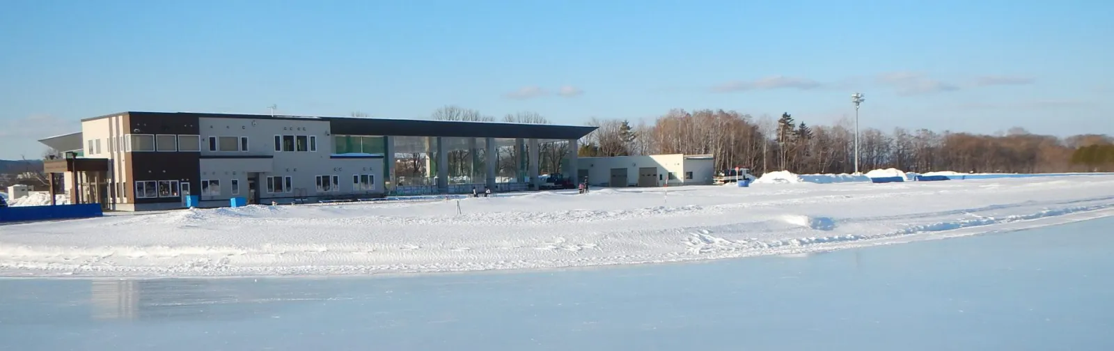 晴れた冬の日に氷面越しに見る、リンクの建物と雪に縁どられたオーバル。 — Kitami Citizens' Skate Rink