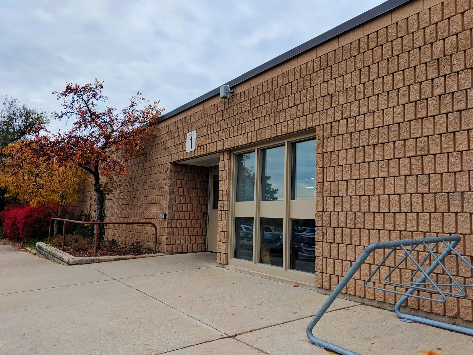 A tan patterned-brick wall of Lions Arena with a numbered entrance door, tall windows and a bike rack in the foreground, under an overcast sky with an autumn tree beside it. — Lions Arena (Kitchener)