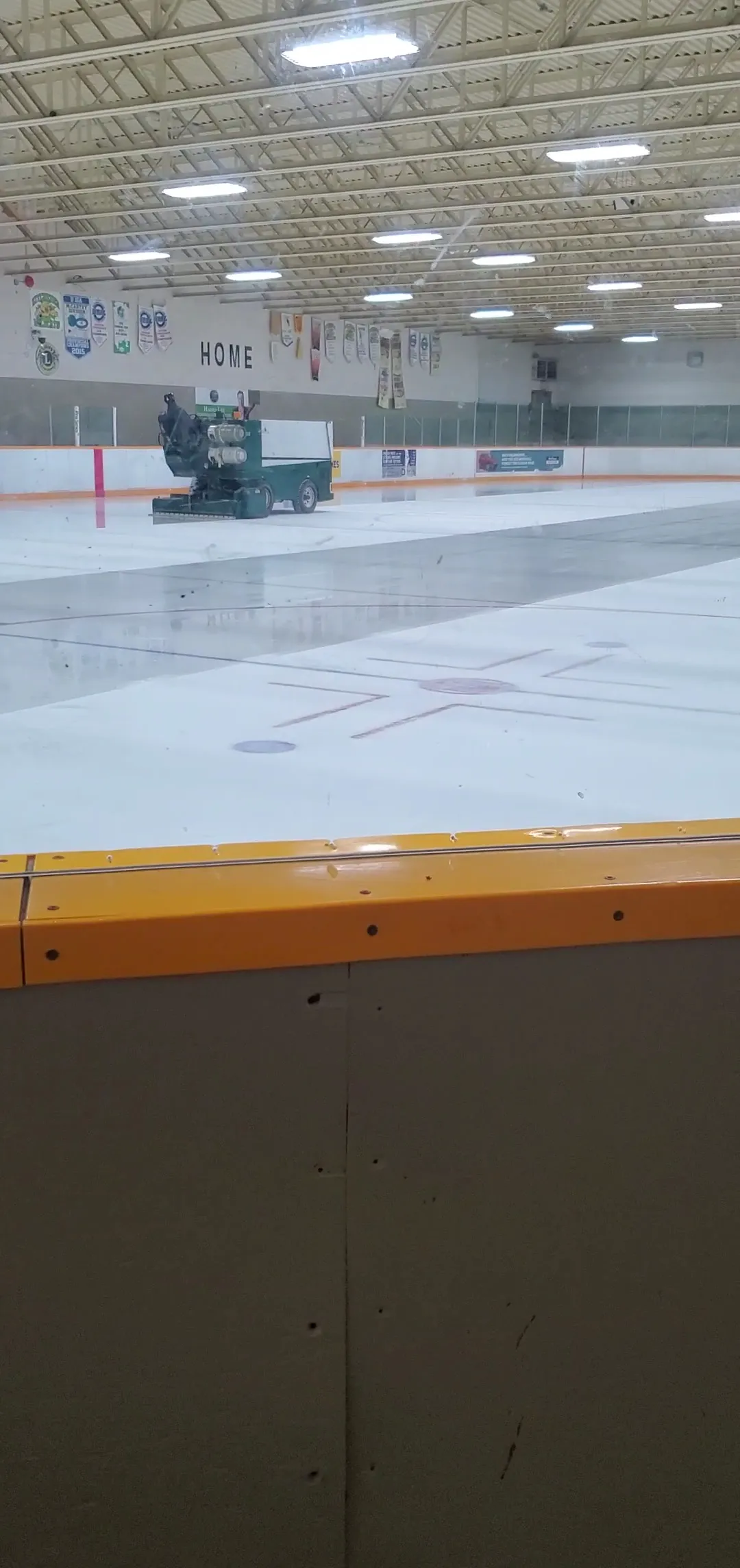 Hielo recién pulido en Lions Arena en Kitchener con la pulidora en el extremo opuesto, visto sobre las vallas con borde naranja bajo el techo de acero — Lions Arena (Kitchener)