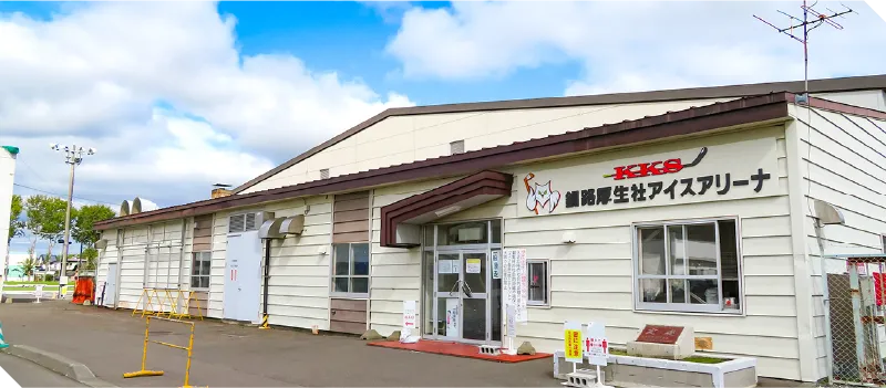 晴れた日のKKS釧路厚生社アイスアリーナの正面。クリーム色の外壁の低い建物で、壁にKKSの文字があり、ガラス張りの入口の前に駐車場が広がっている。 — KKS Kushiro Koseisha Ice Arena