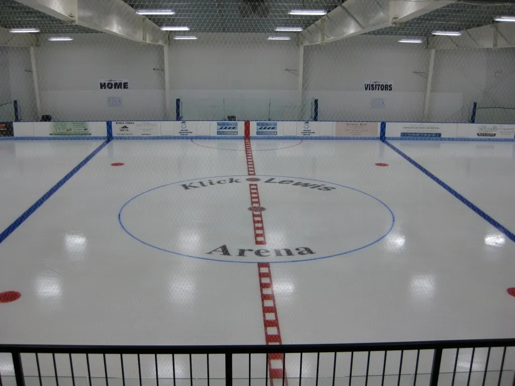 An empty sheet with the arena name at the centre circle, a red centre line and blue lines on white ice under a pale ceiling. — Klick Lewis Arena
