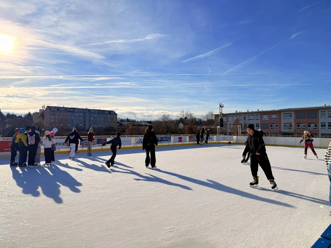 Skaters spread across the open-air rink at Hostivice in bright winter sun, with long shadows on the ice and housing blocks beyond the boards. — Kluziště Hostivice