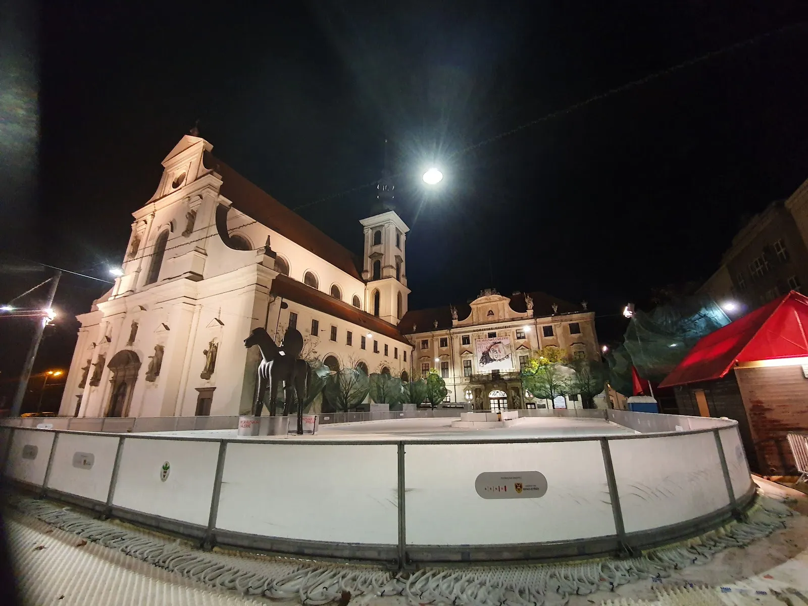 The Moravské náměstí rink in Brno at night, the white boards in front of the church and the Margrave statue — Kluziště na Moraváku Brno