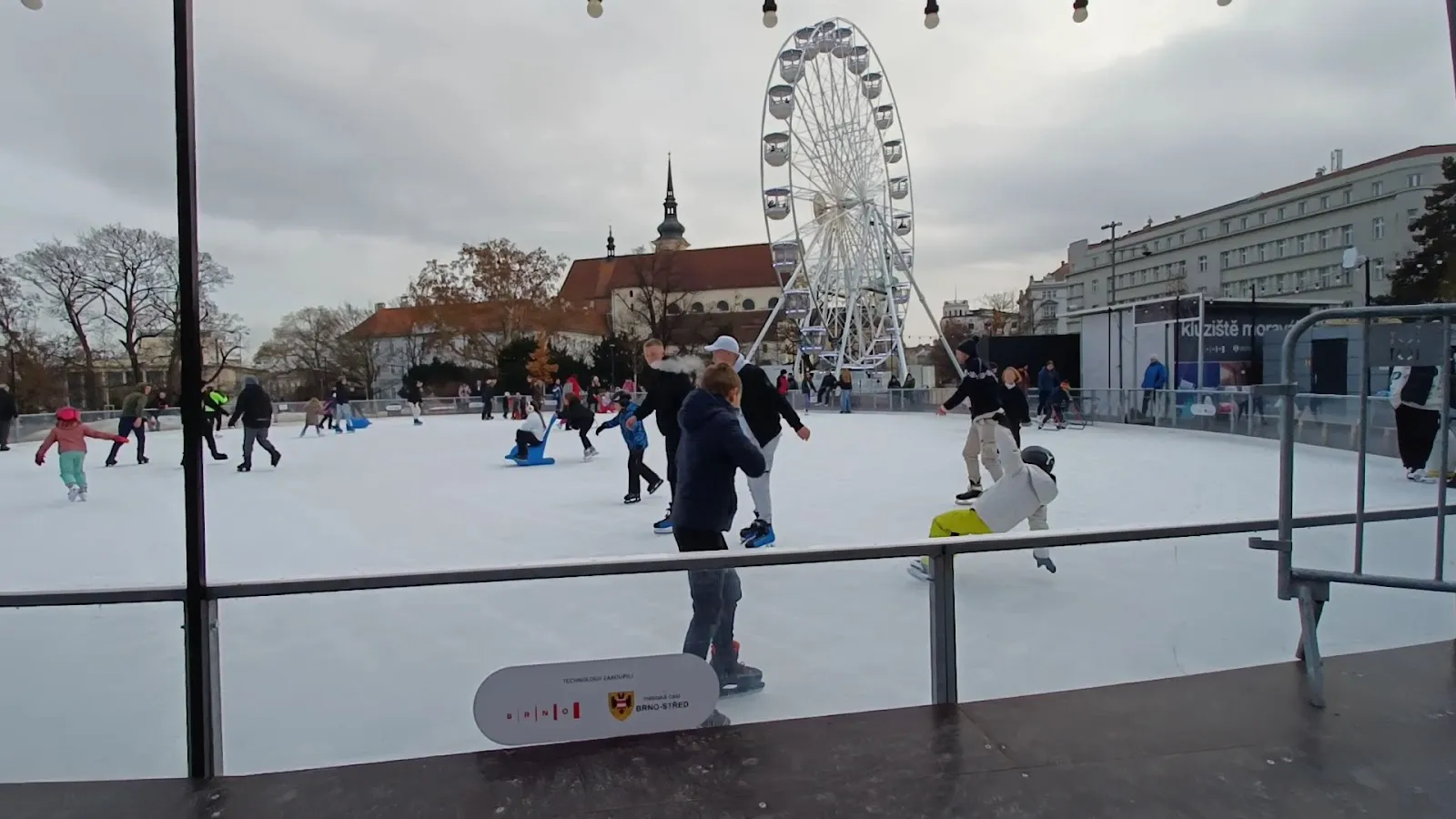 Skaters on the ice of the Moravské náměstí rink in Brno with the Ferris wheel and the old town behind — Kluziště na Moraváku Brno
