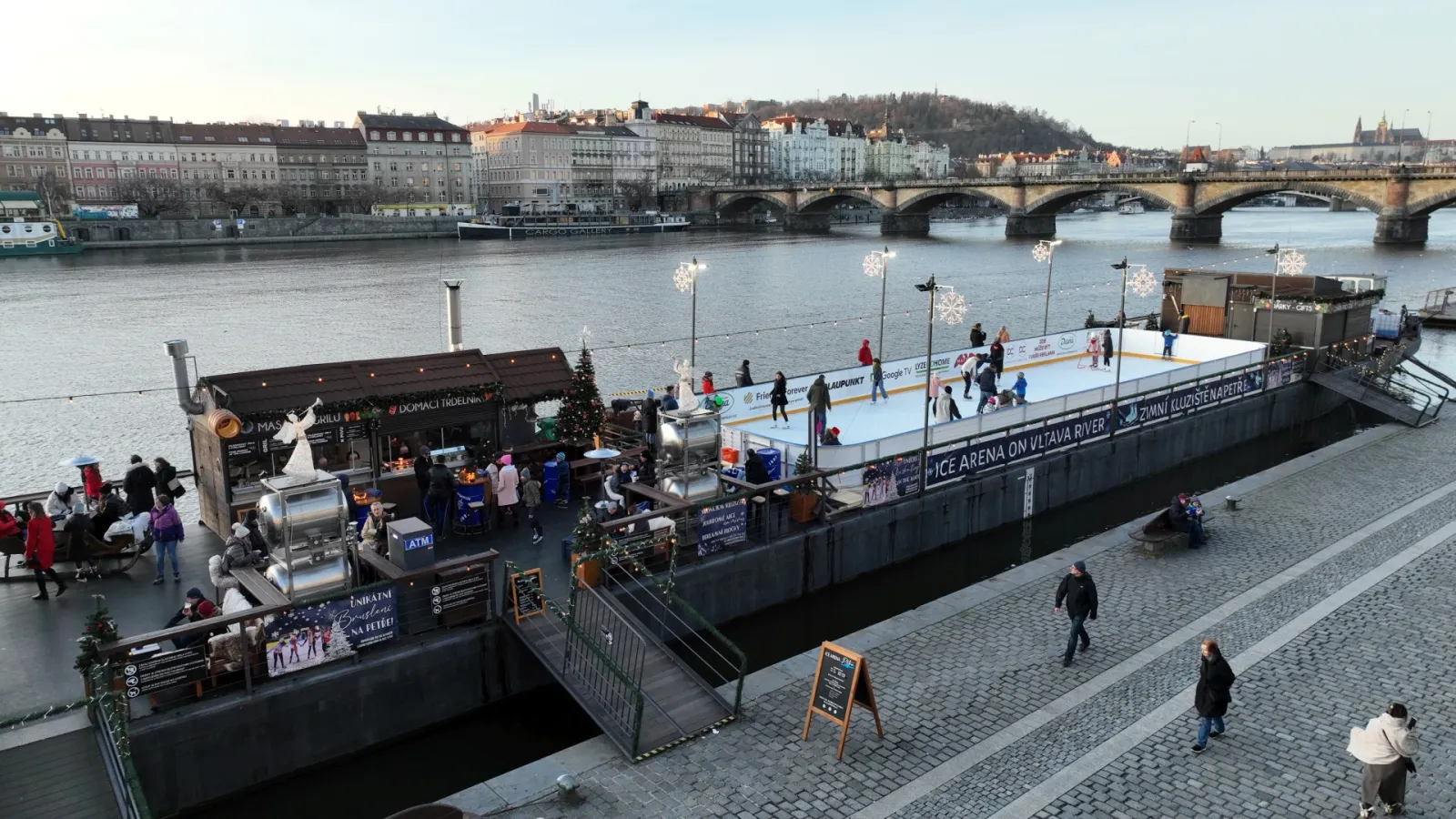 Aerial view of the Petra rink on the Rašín embankment beside the Vltava, with the boarded rink, small huts and Prague's bridges beyond. — Kluziště Petra (Praha 2)