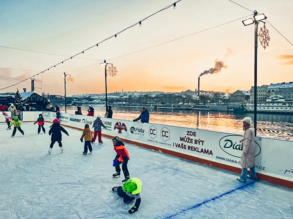 Children skating at sunset on the Petra rink beside the Vltava, with sponsor boards along the rail and a string of lamps overhead. — Kluziště Petra (Praha 2)